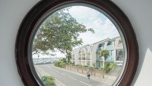A view through a round window with a dark wooden frame, overlooking a street with a building featuring large windows and a tree. - Sea Breeze at Fishermans Dock (Poole)
