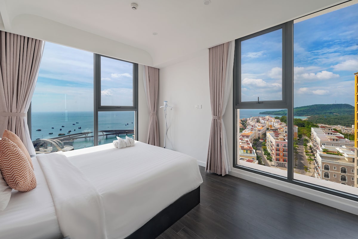 Een moderne hotelkamer met een groot bed, uitzicht op zee en de stad, en een balkon met een hekwerk.
