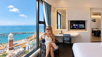 A hotel room with a large window overlooking a coastal cityscape, a bed, a desk with a flat-screen TV, and a seating area with a chair.