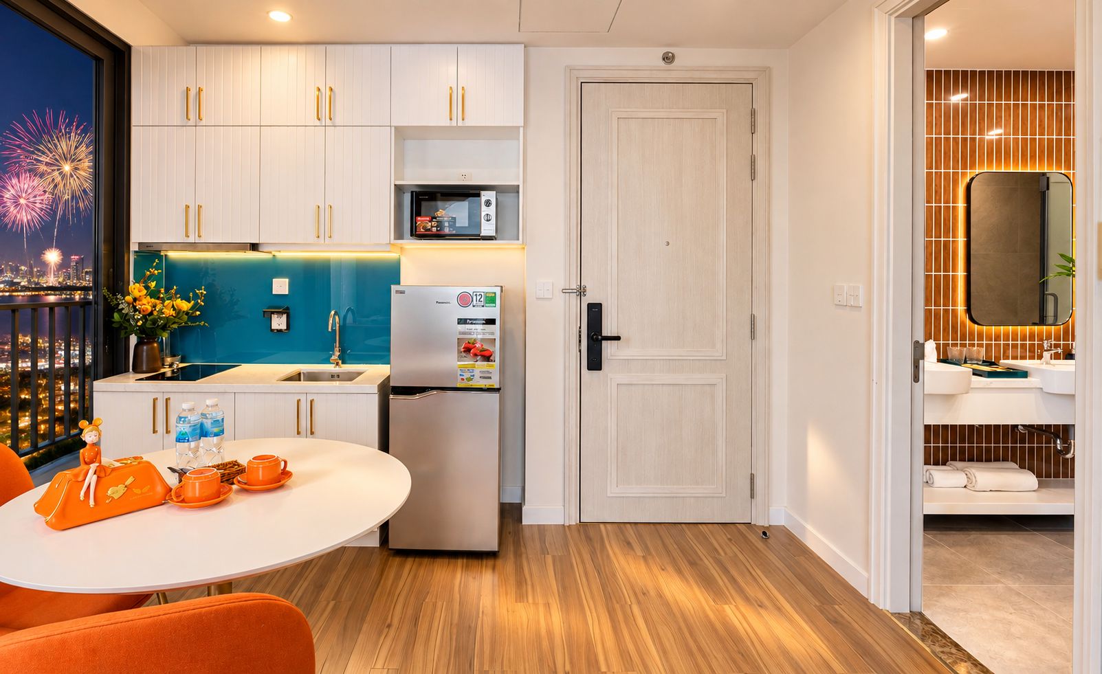 Eine moderne Küche mit weißen Schränken, einem Kühlschrank, einer Mikrowelle und einem Spülbecken. Ein Esstisch mit orangefarbenen Stühlen und eine Blumenvase stehen bereit.