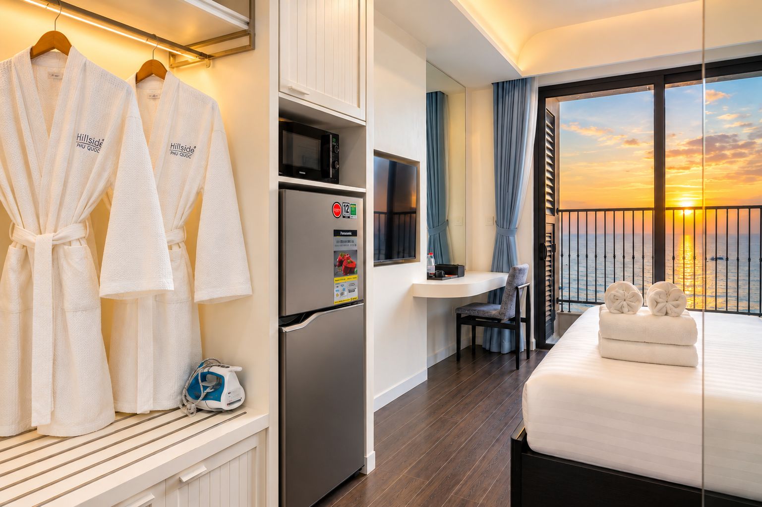 Ein Hotelzimmer mit Bett, Minibar, Kühlschrank, Fernseher und Blick auf den Sonnenuntergang über dem Wasser.