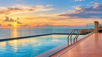 Piscina com vista para o pôr do sol sobre o oceano, deck revestido de azulejos e guarda-corpo de metal.
