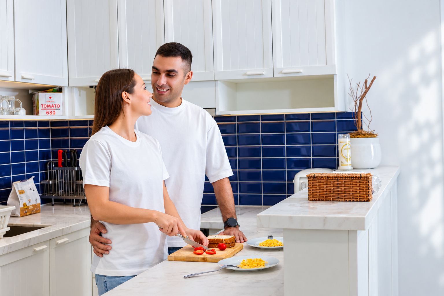 A couple in a kitchen with white cabinets, a blue backsplash, and a basket on the counter.