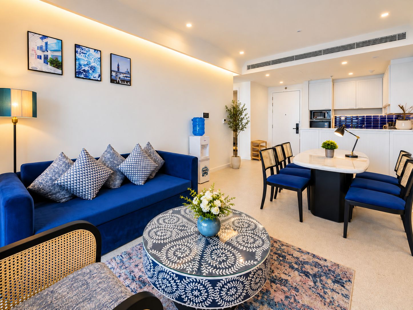 A modern living room with a blue sofa, patterned cushions, a round coffee table with a floral centerpiece, and a dining area with blue chairs and a white table.
