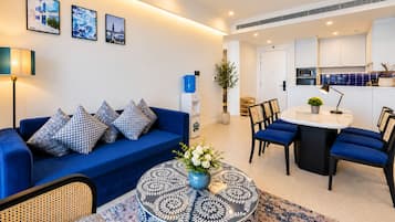 Een moderne woonkamer met een blauwe bank, gestreepte kussens, een ronde salontafel met een bloemstuk, en een eethoek met blauwe stoelen en een witte tafel.