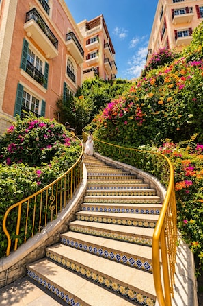 Una escalera con baldosas estampadas, rodeada de abundante vegetación y flores coloridas, que conduce a un edificio con balcones.