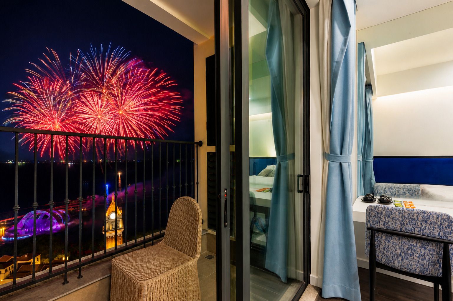 A balcony with a view of fireworks, a wicker chair, and a bed with a blue headboard.