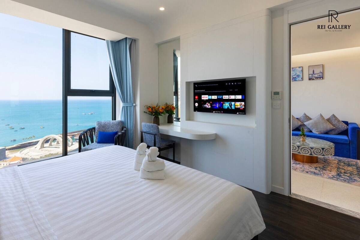 Une chambre d’hôtel dotée d’un grand lit, d’un téléviseur fixé au mur, d’un coin salon avec un canapé et des fauteuils, et offrant une vue sur la mer.