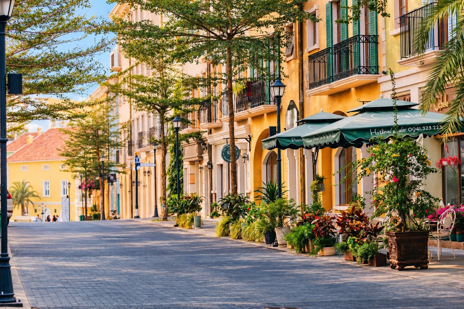 Una calle flanqueada por edificios amarillos, con mesas al aire libre y sombrillas, y plantas en macetas.