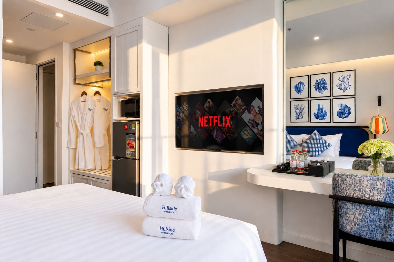 A hotel room with a bed, a TV displaying the Netflix logo, a minibar, and a seating area with a vase of flowers.