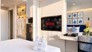 Une chambre d’hôtel avec un lit, un téléviseur affichant le logo de Netflix, un minibar et un coin salon agrémenté d’un vase de fleurs.