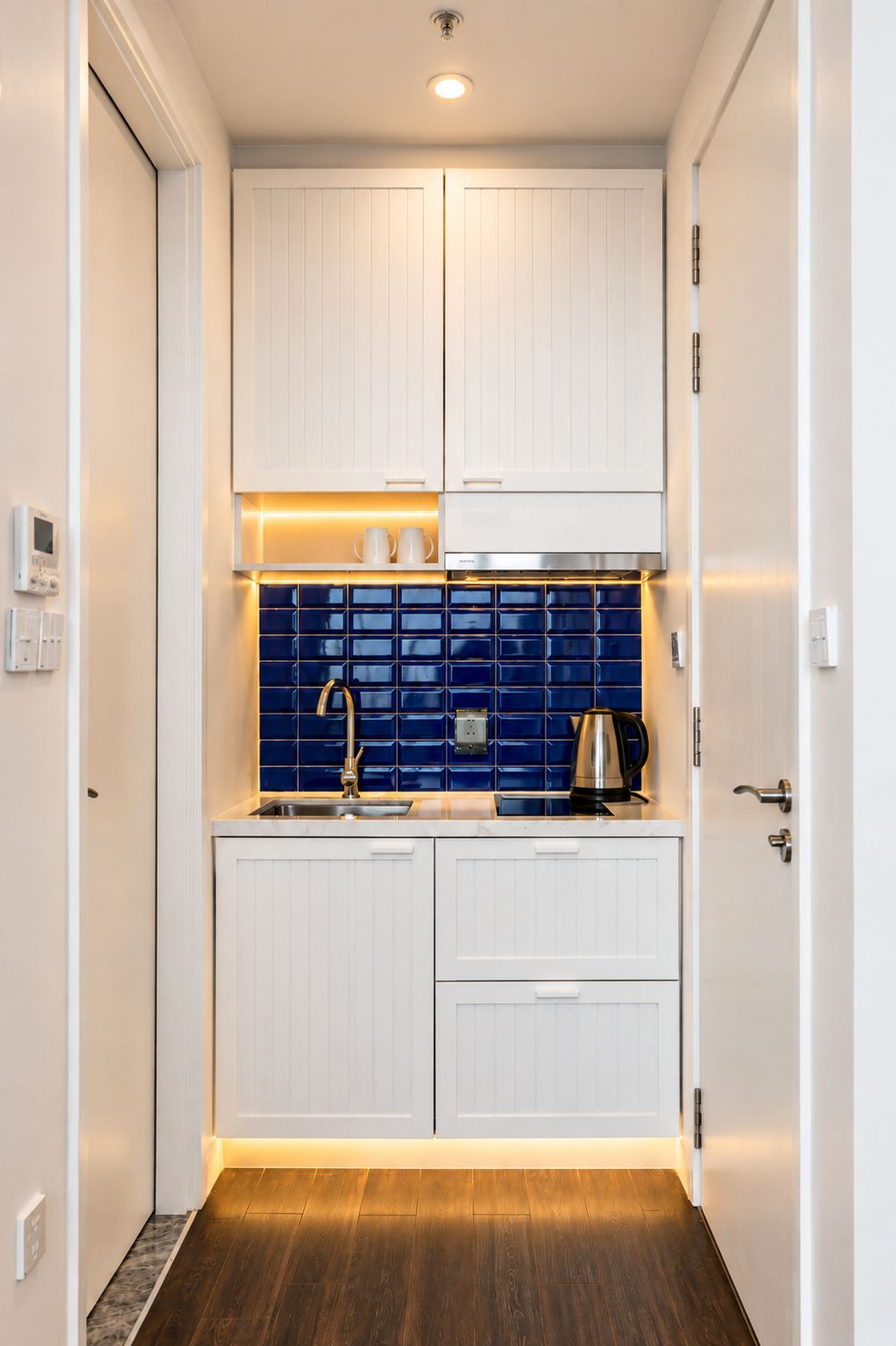 A compact kitchen with white cabinetry, a blue tiled backsplash, and a wooden floor.