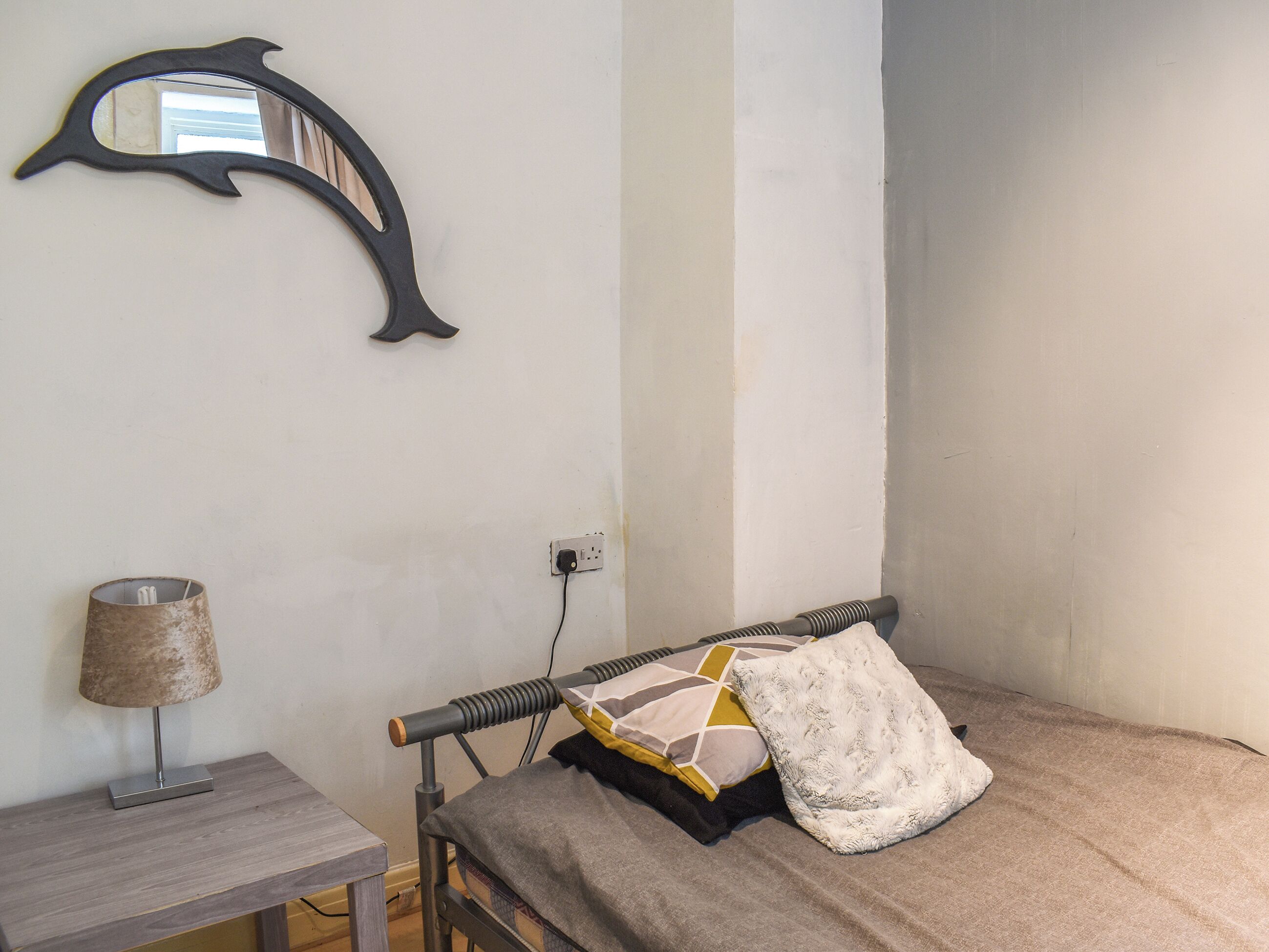 A bedroom with a bed, a bedside table with a lamp, a wall-mounted mirror, and a power outlet.
