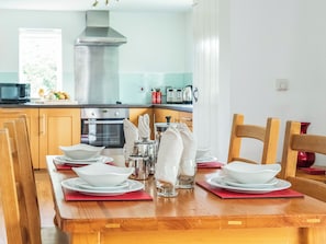 A dining table set with white plates, bowls, and glasses, surrounded by wooden chairs in a kitchen with wooden cabinets and a stainless steel range hood. - 1 bedroom accommodation in Crackington Haven (Crackington Haven)