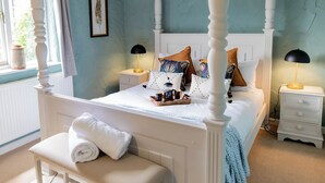 Ein Schlafzimmer mit einem Himmelbett, weißer Bettwäsche und vielen Kissen. Ein Nachttisch mit Lampe, ein Beistelltisch und ein Fenster mit Vorhängen sind ebenfalls vorhanden. Ein zusammengerolltes Handtuch liegt auf einem Stuhl vor dem Bett.