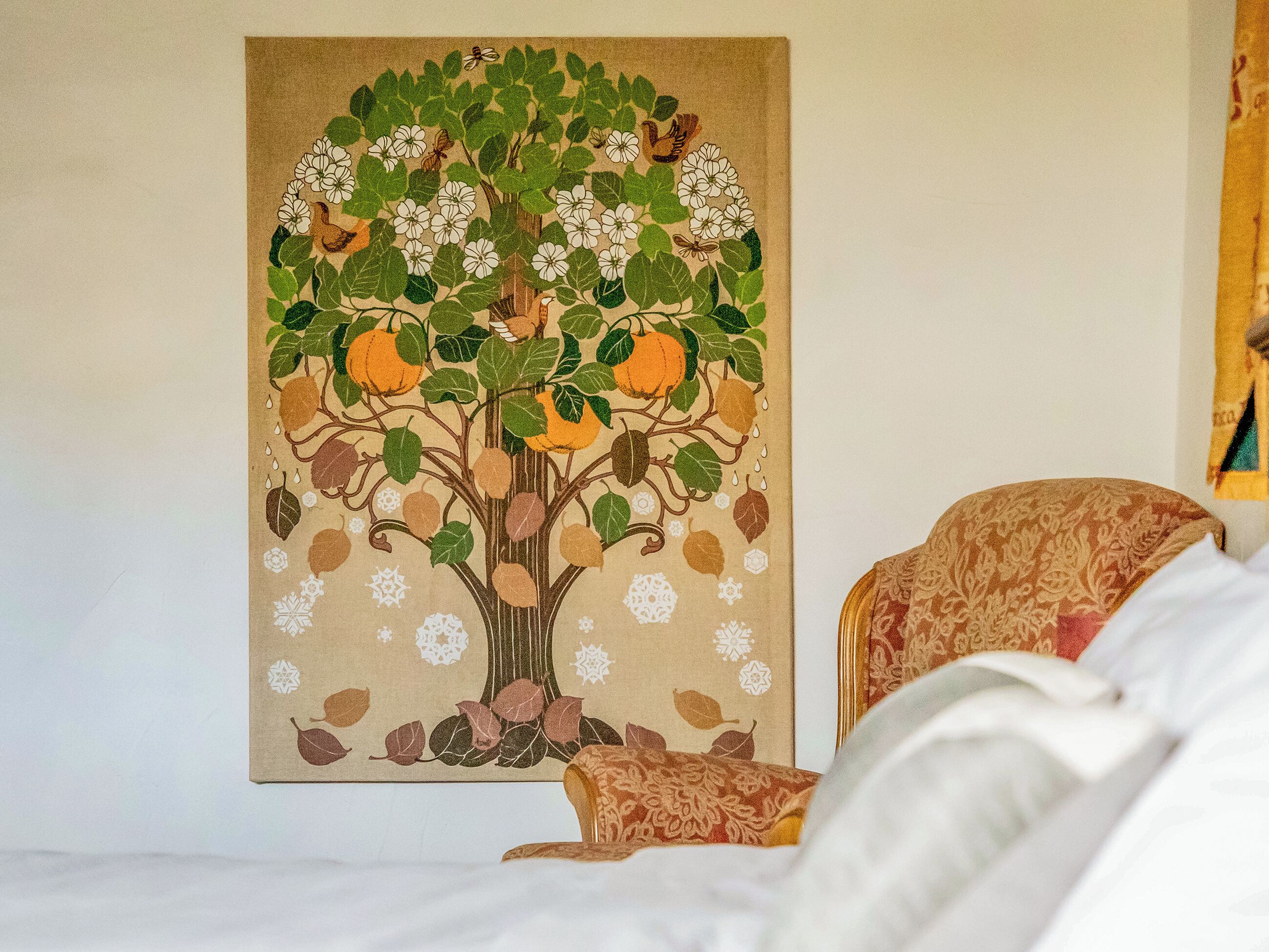 A room with a bed, a chair, and a large wall tapestry depicting a tree with leaves and flowers.