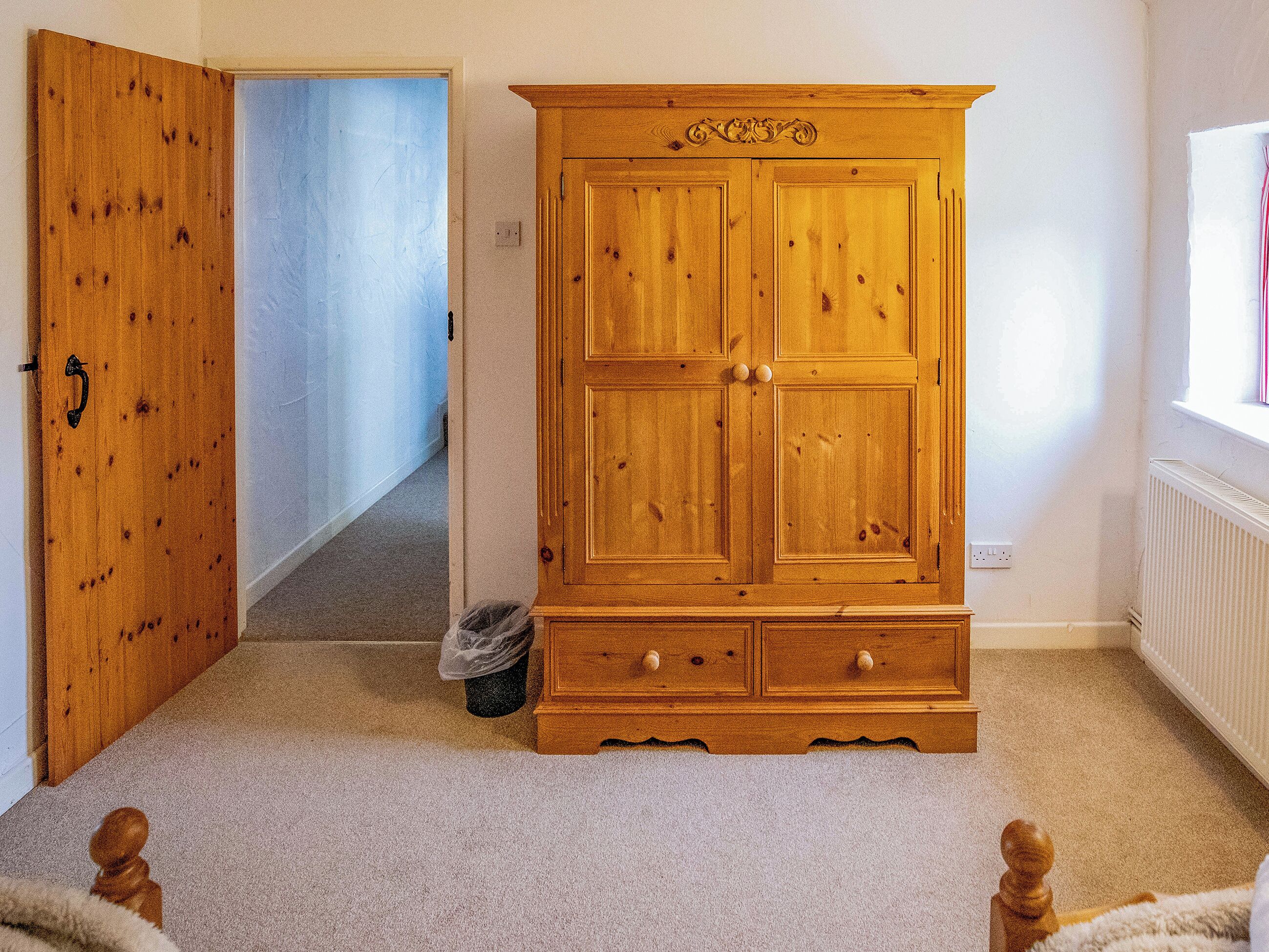 A wooden wardrobe with two doors and a lower shelf, a carpeted floor, and a hallway leading to another room.