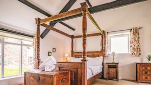 A four-poster bed with a wooden headboard, a nightstand with a lamp, a wooden chest of drawers, and a window with curtains. - 1 bedroom accommodation in Crackington Haven (Crackington Haven)