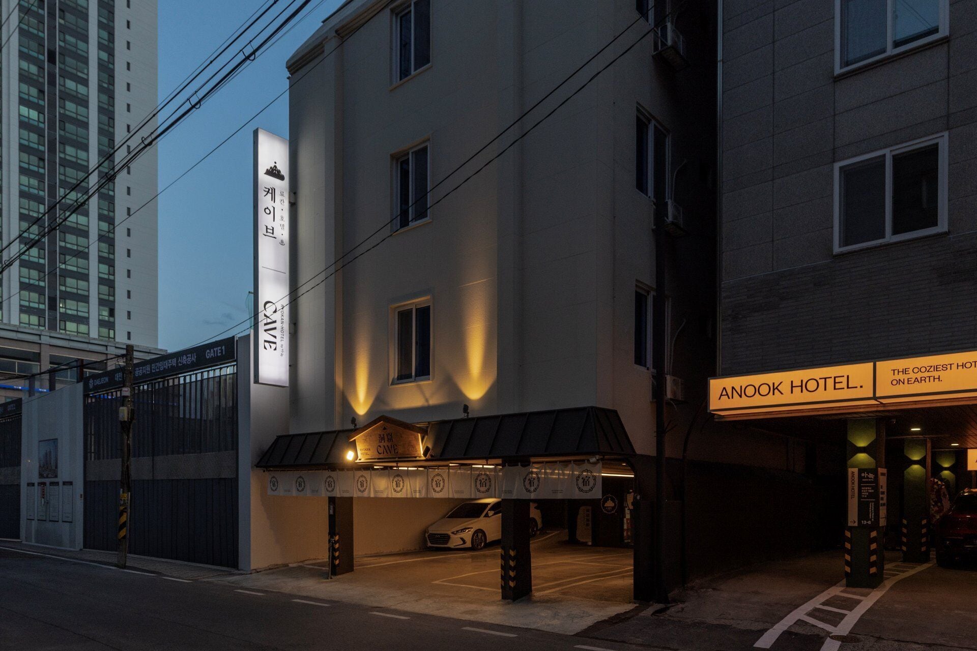 A hotel entrance with a sign reading 'ANOOK HOTEL' and a parking area with a white car.