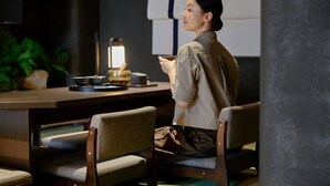 Iemand zit aan een houten tafel met een laptop, een lamp en een kom. Er staan stoelen om de tafel heen en een plantje op de achtergrond.