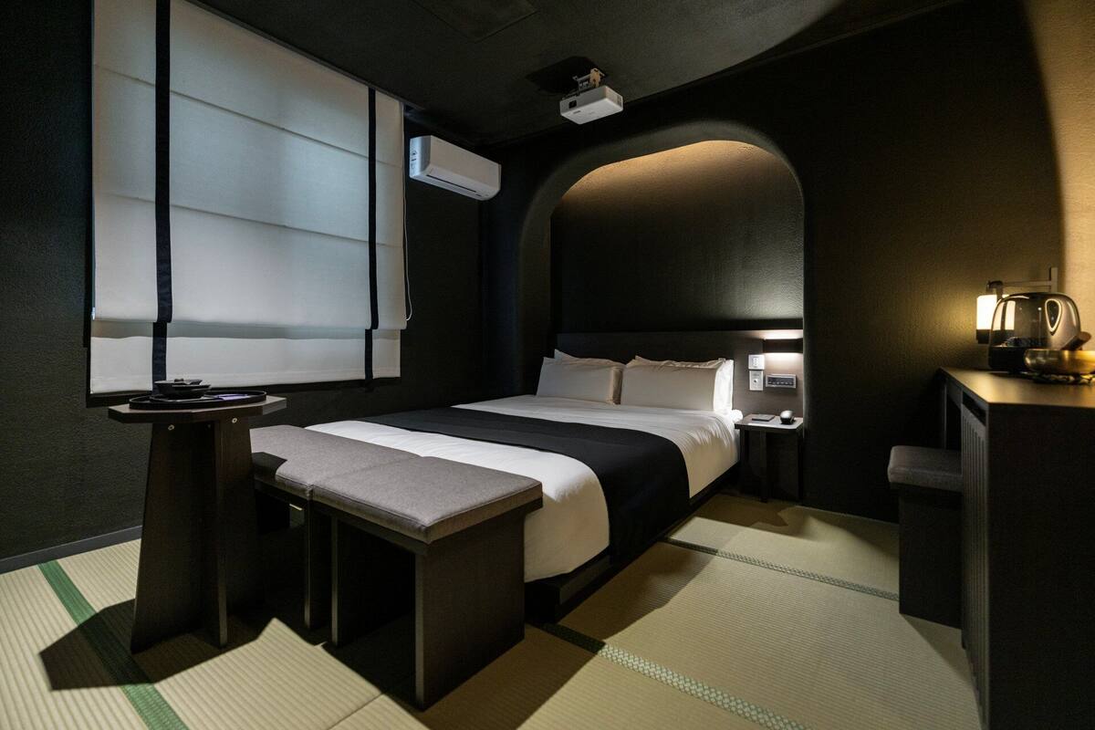 Habitación de hotel con cama, mesita de noche, lámpara y un proyector en el techo.