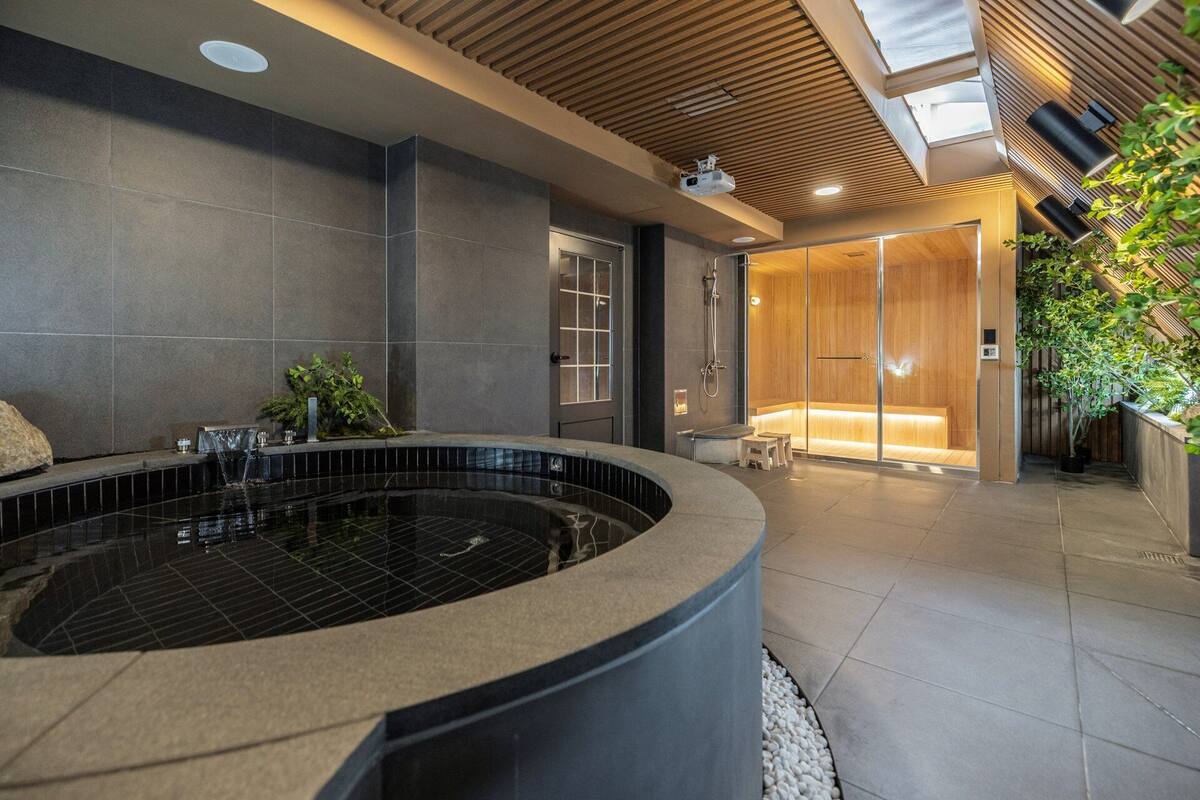 Un baño moderno con una bañera de hidromasaje grande y circular, un cerramiento de ducha de vidrio y una zona de estar con un banco de madera.