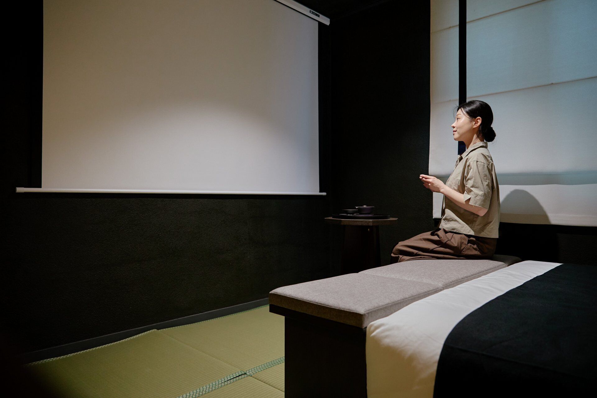 Una persona sentada en un esterilla de tatami en una habitación con una pantalla de proyector, una mesita con un cuenco y una cama con una colcha blanco y negro.