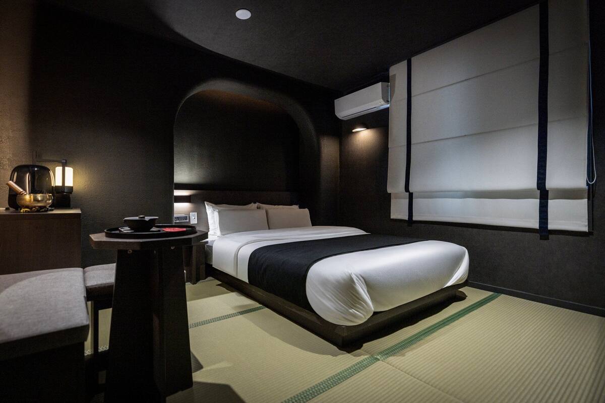 Una habitación de hotel moderna con una cama, una mesita de noche con lámpara, una silla y aire acondicionado instalado en la pared.