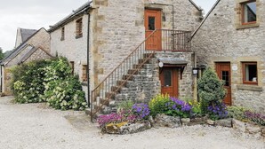 Edifici de pedra amb una escala de fusta que condueix a una porta, envoltat de grava i flors de colors.