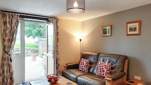Ein Wohnzimmer mit einer Ledersofa, einem Holztisch, einer Stehlampe und Blick auf den Garten durch eine Glastür.