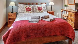 Ein ordentlich bezogenes Bett mit roter Decke, zwei Kissen mit Blumenmuster und einem hölzernen Kopfteil. Zwei Nachttische mit Lampen, eine Holzkommode und ein Spiegel an der Wand sind ebenfalls zu sehen.