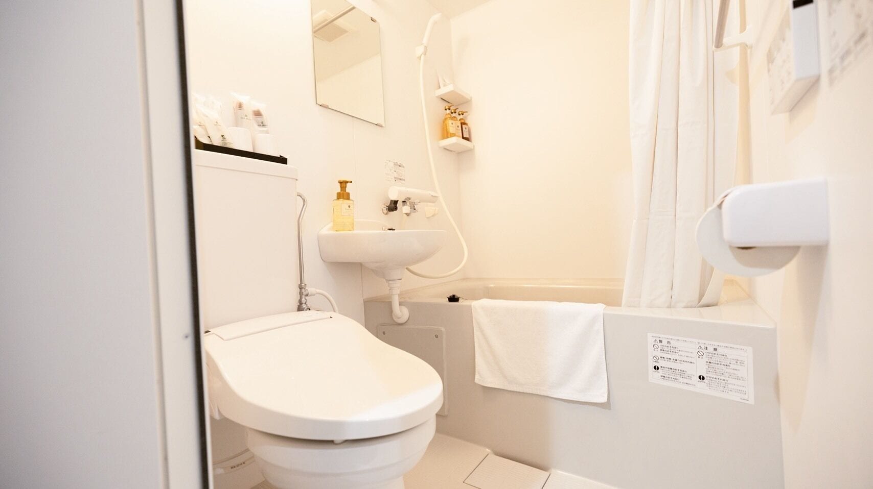 Une petite salle de bain propre, équipée de toilettes, d’un lavabo et d’une douche. Une serviette est accrochée au mur et un distributeur de savon est posé sur le lavabo.
