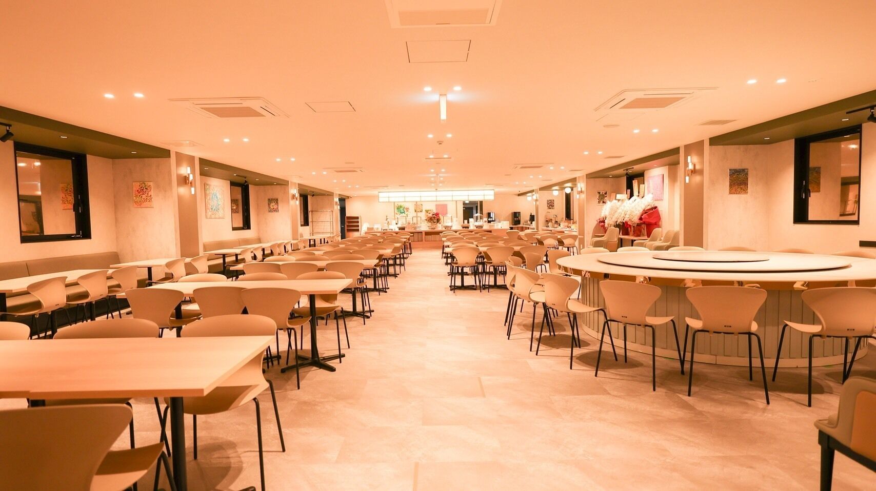 A spacious hotel dining area with multiple tables and chairs, a central round table, and a long counter with a display of items.