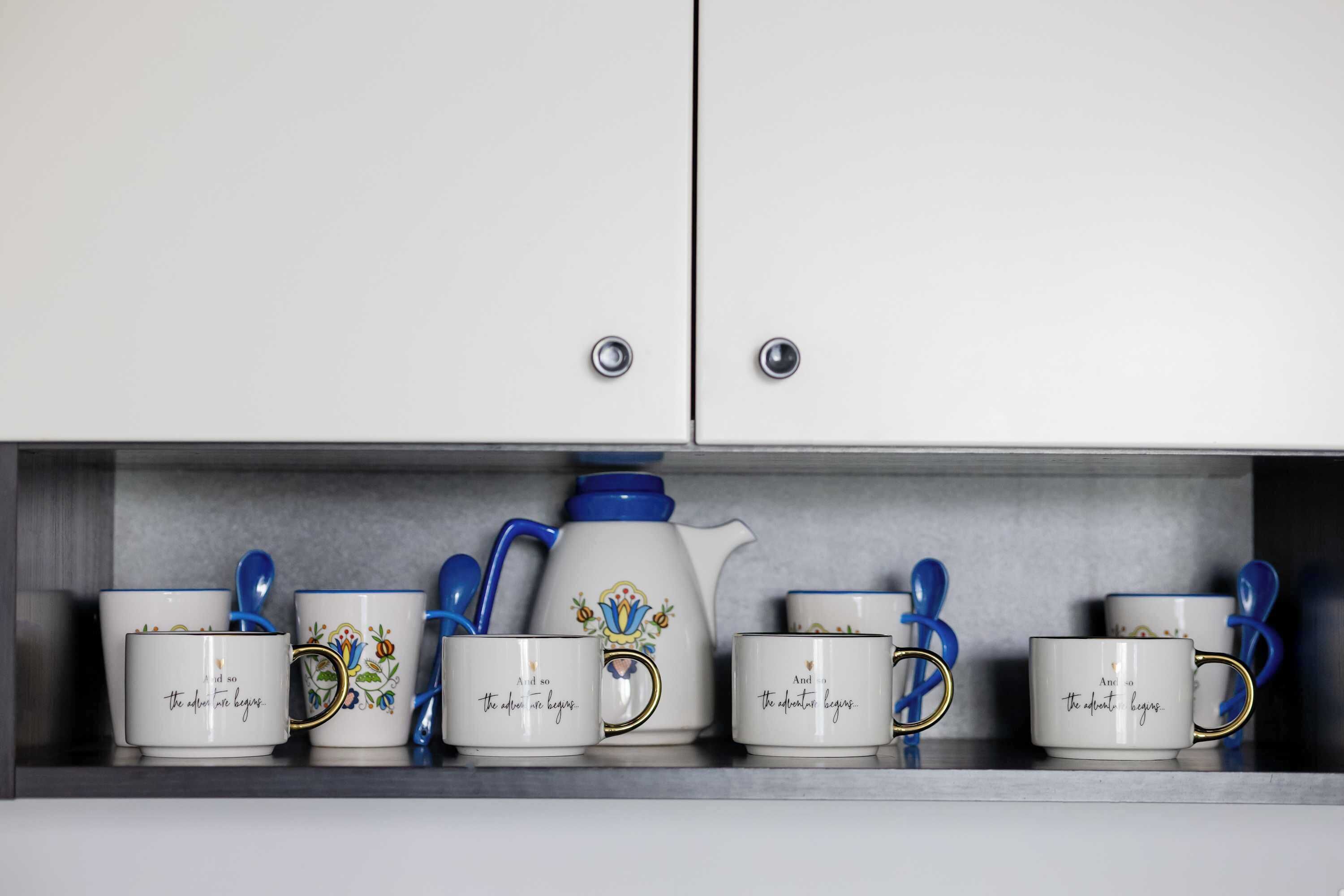 Een verzameling witte mokken met blauwe handgrepen en een blauwe theepot op een plank.