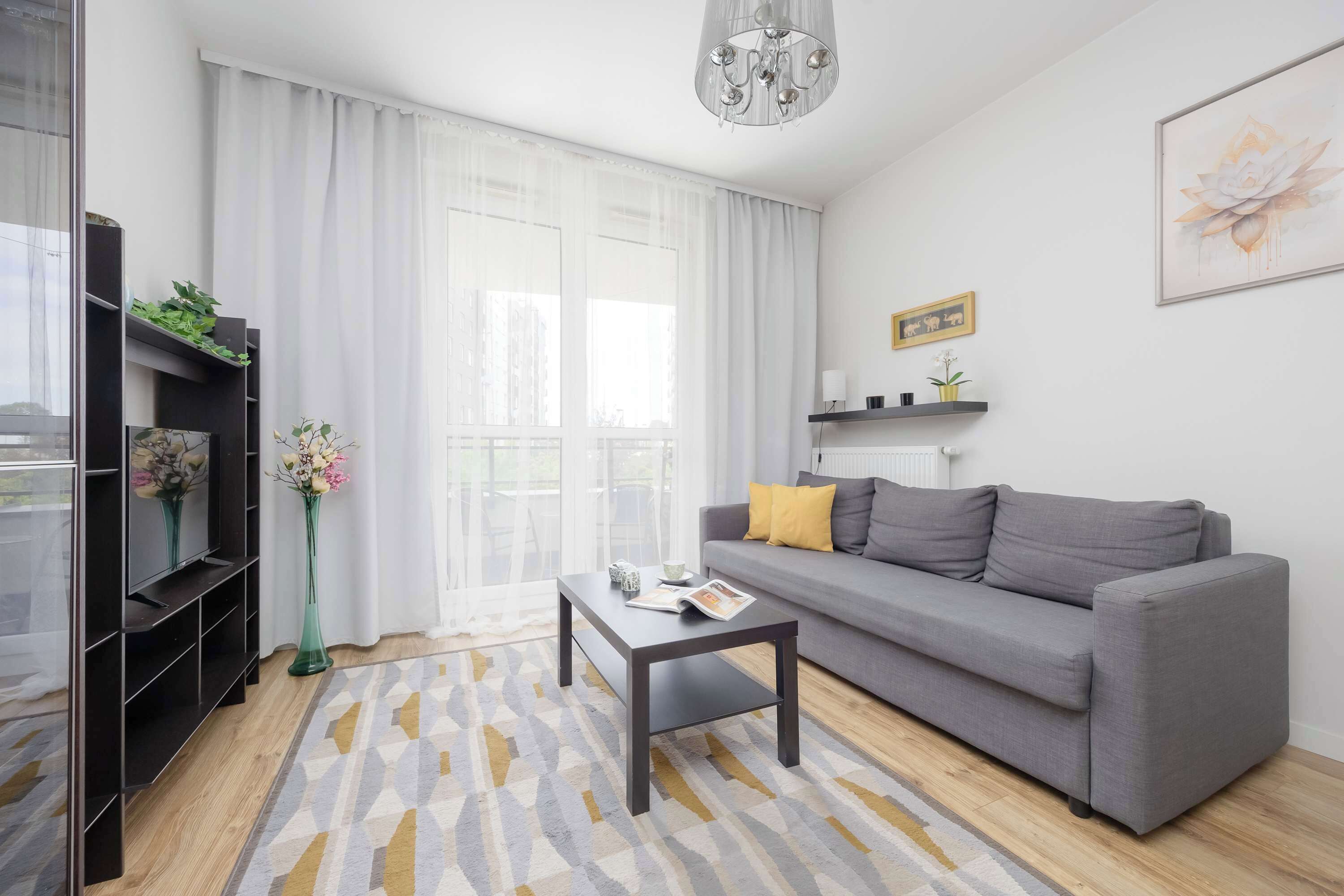Een moderne woonkamer met een grijze bank, een salontafel, een televisie op een zwart meubelstuk en een groot raam met dunne gordijnen.