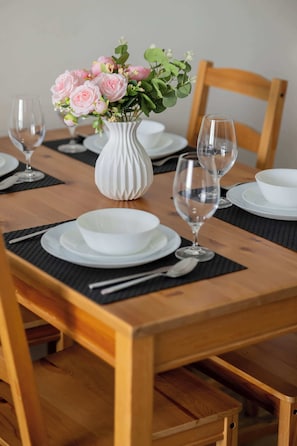 Ein Holztisch ist mit weißen Tellern, Schüsseln und Besteck gedeckt, dazu stehen eine Vase mit rosa Blumen und zwei Weingläser.