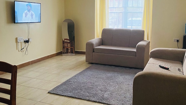 Olohuoneessa on beigenvärinen sohva, harmaa matto, seinälle asennettu televisio ja ikkuna, jossa on verhot.