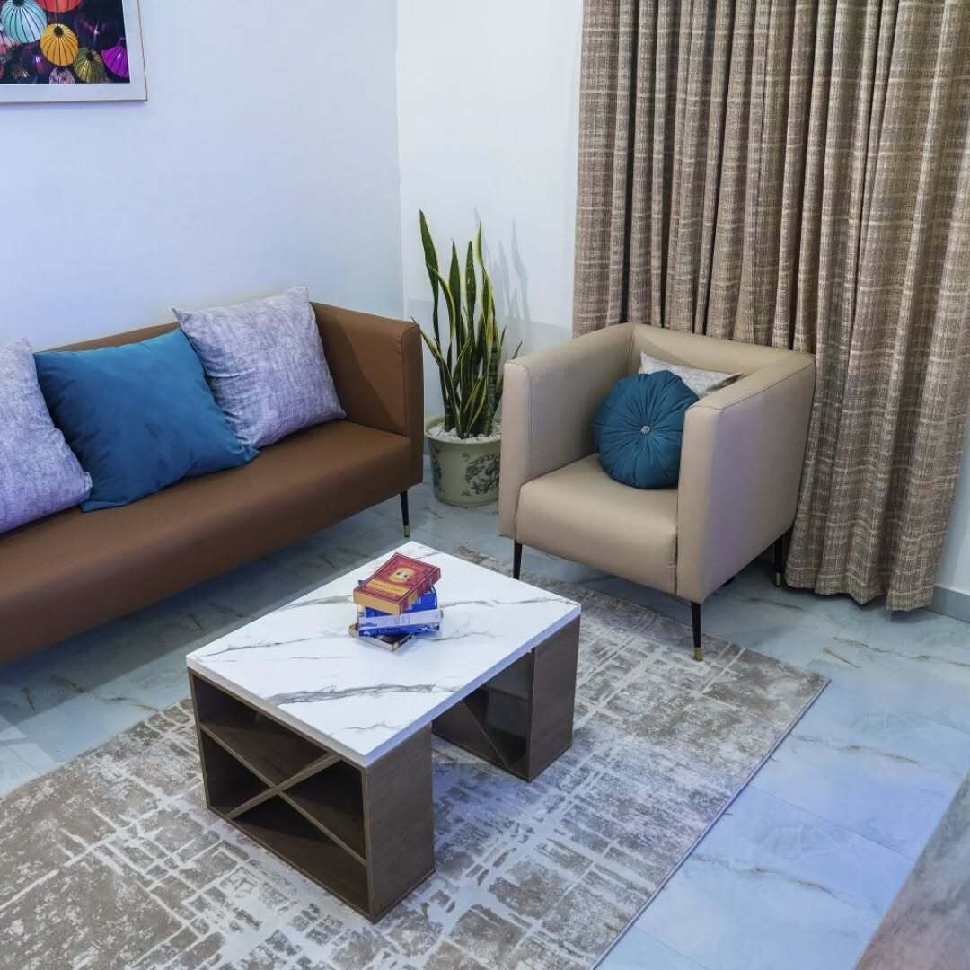 A living room with a brown sofa, a beige armchair, a marble coffee table, and a potted plant.