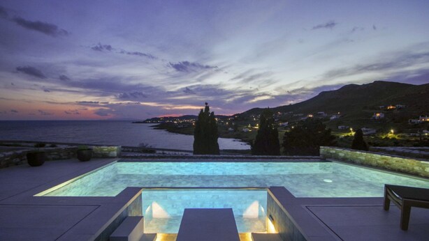 An infinity pool with a view of the sea and a town at dusk.