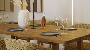 A wooden dining table with four plates, two wine glasses, a bottle of wine, and a vase with dried plants.