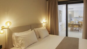 A hotel room with a bed, two bedside lamps, a view of the dining area through a glass door.