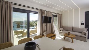 A modern living room with a sofa, a TV mounted on the wall, a dining table with chairs, and a view of the sea through large windows. - Villa Marenthe | 3 BDR | Sea Views | Pool | Syros (Syros)