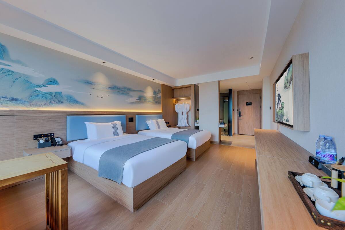 Ein modernes Hotelzimmer mit zwei Betten, einem Holztisch, einem großen Wandbild und einem durchgegangene Tür erkennbaren Badezimmer.