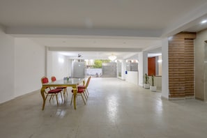 A spacious dining area with a long table and red chairs, a kitchen in the background, and a wooden accent wall. - Yashobhoomi Suites near Yashobhoomi Convention Centre (New Delhi)