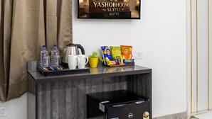 A hotel reception area with a mounted TV displaying the hotel's name, a coffee machine, bottled water, snacks, and a black refrigerator. - Yashobhoomi Suites near Yashobhoomi Convention Centre (New Delhi)
