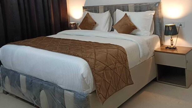 A neatly made bed with a headboard, a bedside table with a lamp, and an air conditioning unit above the bed. - Yashobhoomi Suites near Yashobhoomi Convention Centre (New Delhi)
