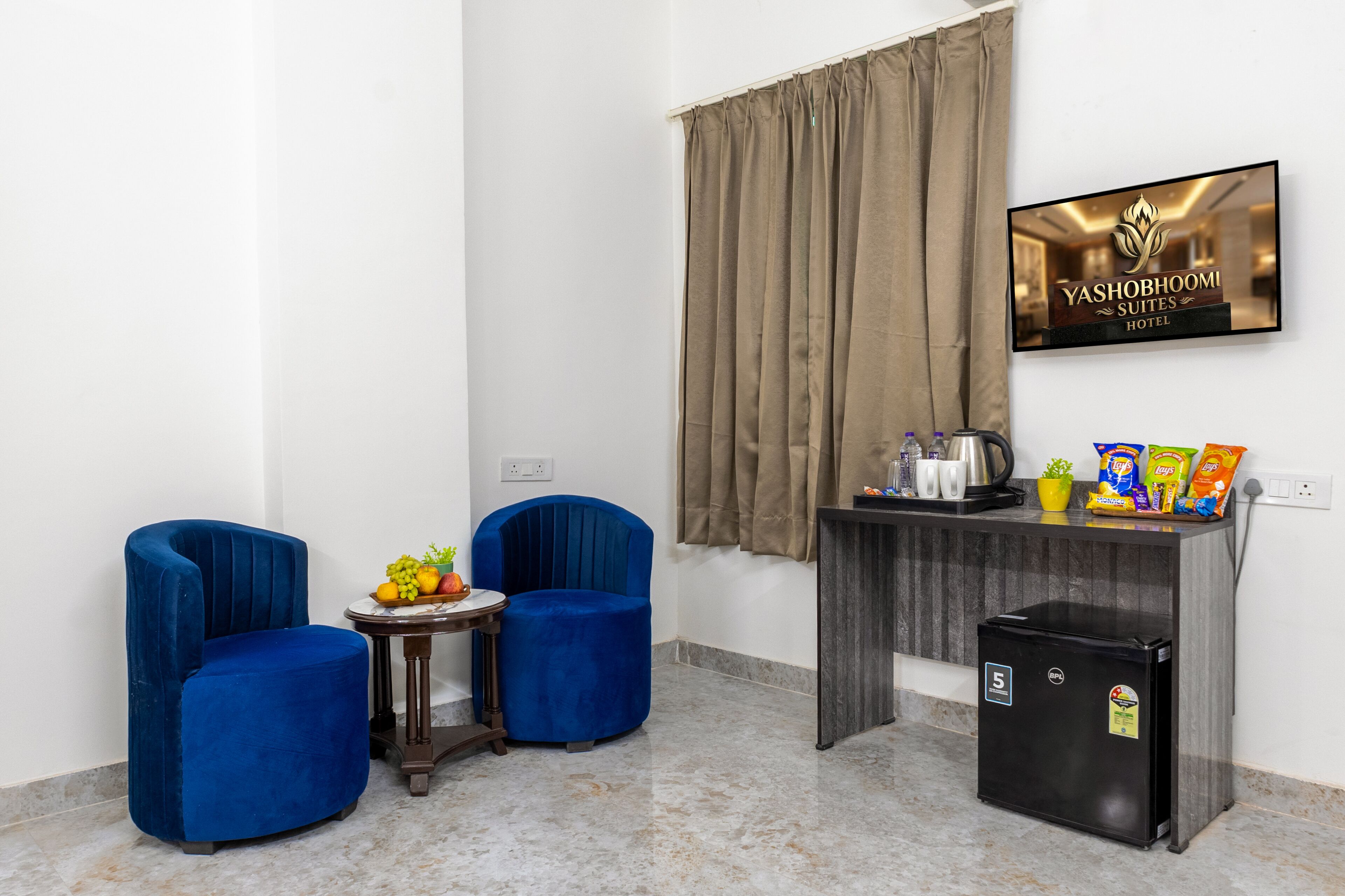 A room with two blue armchairs, a small round table with fruit, a mini fridge, a TV displaying the hotel name, and a curtain.