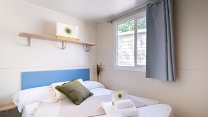A neatly arranged hotel room with a bed, a window with curtains, a shelf with a sign, and a vase with dried flowers. - Friendly Mobile Home in Rimini Family Village (Rimini)