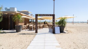 A beachside restaurant named 'Dolcevita 11' with outdoor seating, a clear sky, and a sandy path leading to the entrance. - Friendly Mobile Home in Rimini Family Village (Rimini)