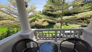 Un balcone con sedie in vimini e un tavolino, affacciato su un paesaggio verde con alberi e un recinto bianco.